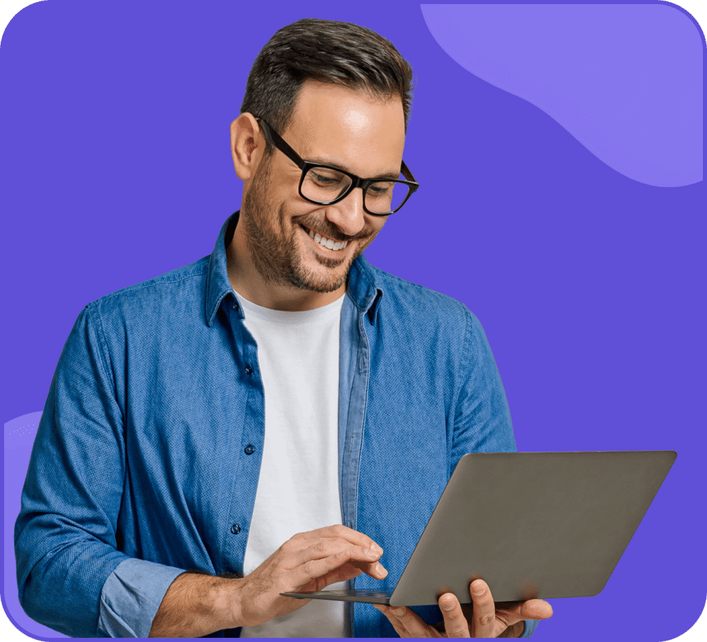 Un homme souriant avec des lunettes, portant une chemise en jean sur un t-shirt blanc, se tient devant un fond violet tout en utilisant un ordinateur portable.