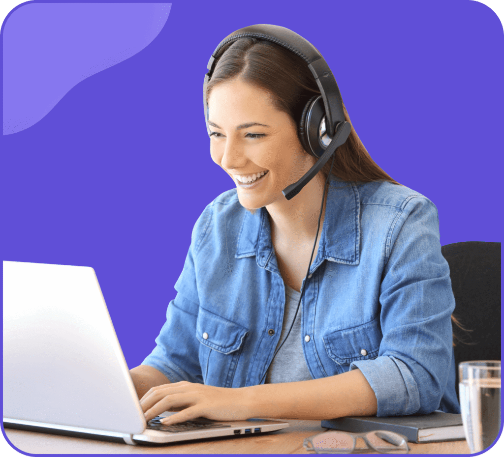 Une personne souriante aux cheveux longs, portant un casque, est assise à un bureau et tape sur un ordinateur portable. Elle porte une chemise en jean bleue, sur un fond violet uni.