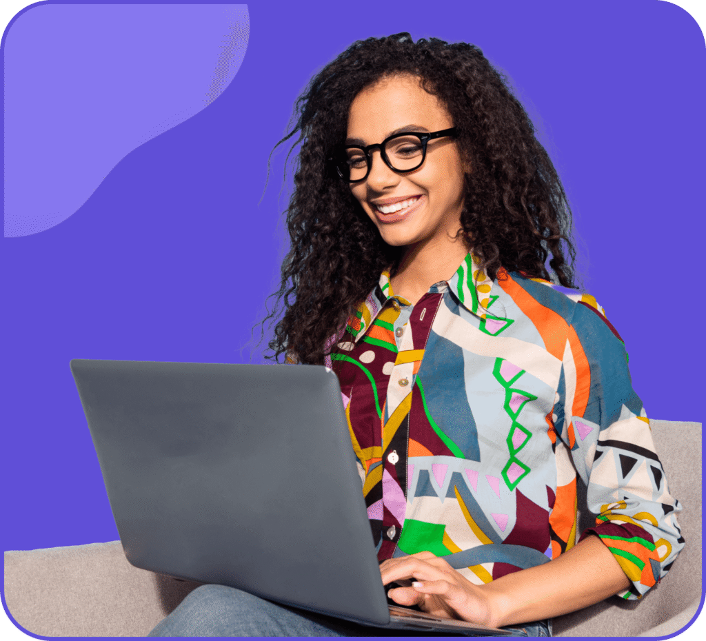 Une femme souriante aux cheveux bouclés et aux lunettes, portant une chemise à motifs colorés, est assise sur un canapé et utilise un ordinateur portable sur un fond violet.